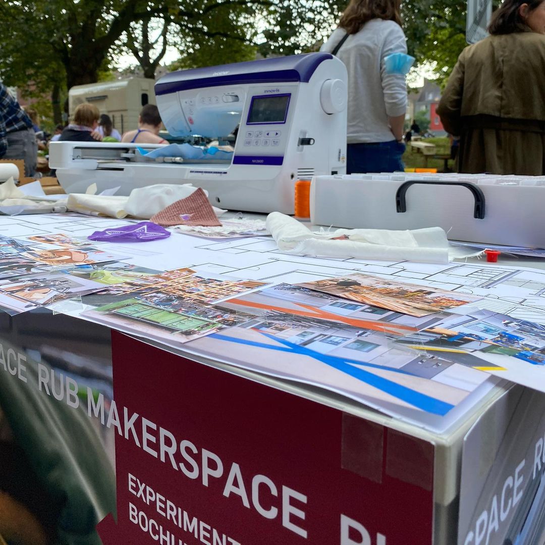 Nahaufnahme unseres Tischs. Im Vordergrund: Bilder des Makerspaces im Aufbau. Dahinter: unsere Stickmaschine.